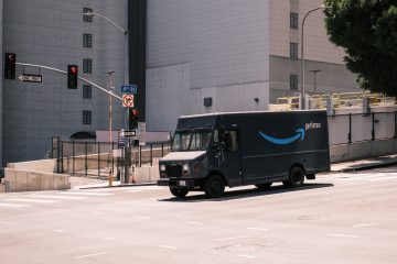 An amazon prime delivery truck drives through an intersection. The truck is dark and slightly ominous while the surroundings are a lighter cityscape.